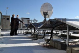 町役場庁舎屋上の太陽光パネル前に立つ小松町長（左）。冬季は雪の反射を取り入れられるよう両面に発電パネルを備える。奥に立つのは蓄電池