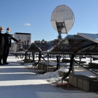 町役場庁舎屋上の太陽光パネル前に立つ小松町長（左）。冬季は雪の反射を取り入れられるよう両面に発電パネルを備える。奥に立つのは蓄電池