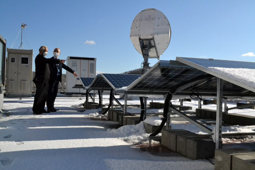町役場庁舎屋上の太陽光パネル前に立つ小松町長(左)。冬季は雪の反射を取り入れられるよう両面に発電パネルを備える。奥に立つのは蓄電池